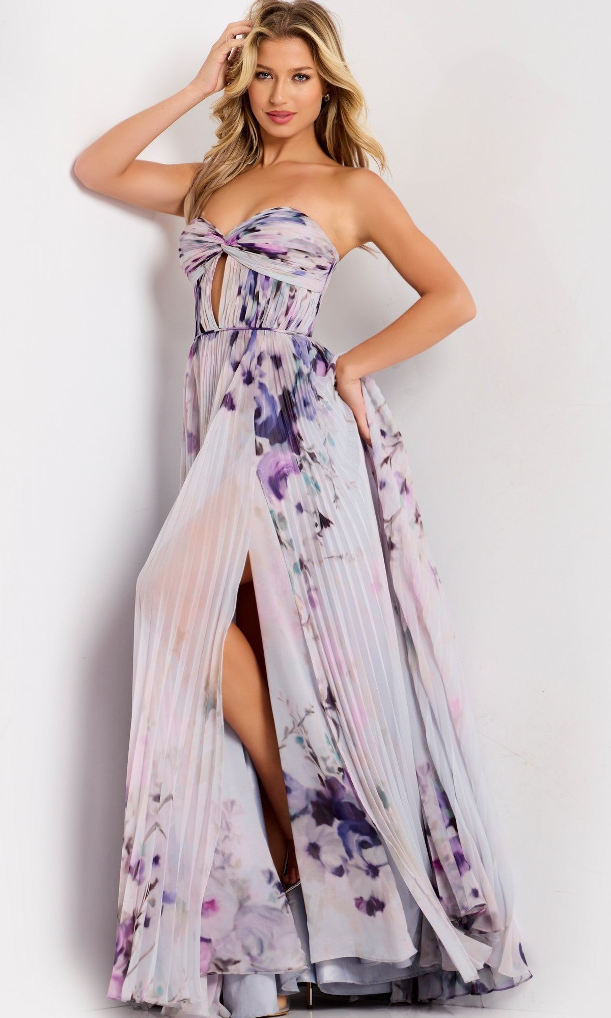 A woman models the Jovani 42564, a strapless long prom dress with a floral print, pleated skirt, front slit, and keyhole cutout, standing against a plain light background.